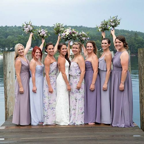 A group of eight women, including one in a white dress, pose on a dock by a lake. Most wear lavender dresses and three hold bouquets aloft, smiling.