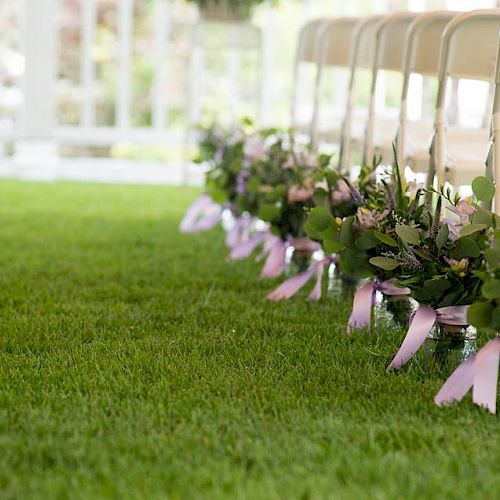 The image shows a row of chairs on a grassy lawn, decorated with floral arrangements tied with pink ribbons, likely set up for an outdoor event.