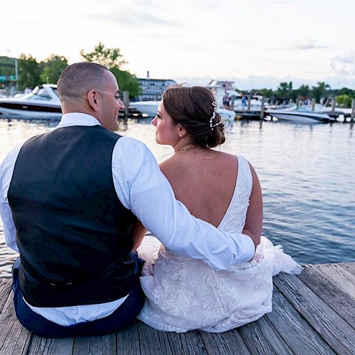 A couple sits on a dock, dressed in wedding attire, gazing at the boats and serene water in the background, sharing a quiet, intimate moment.