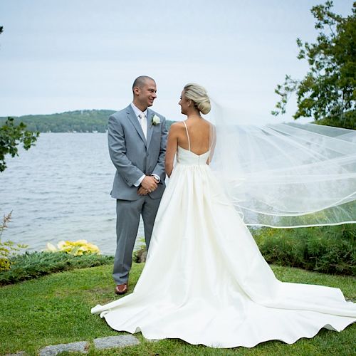 A bride and groom stand together near a lake, with the bride's veil flowing in the wind and a scenic backdrop of trees and water visible.