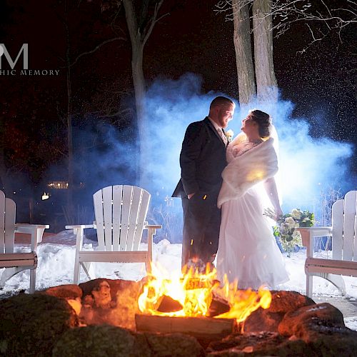 A couple in wedding attire stand near a fire pit surrounded by snow and Adirondack chairs, with a misty backdrop and trees illuminated.