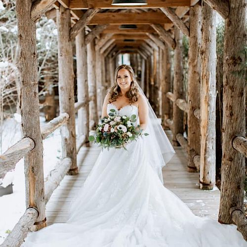 A bride in a white wedding dress stands on a rustic wooden bridge, holding a bouquet of flowers, surrounded by wooden beams and trees.