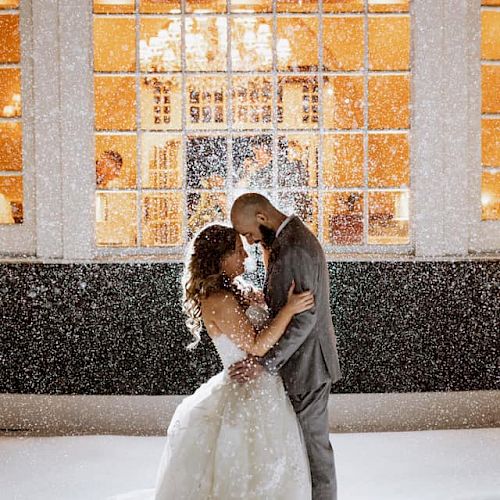 A couple in wedding attire embraces in a snowy scene, with a large window and warm indoor lights in the background.