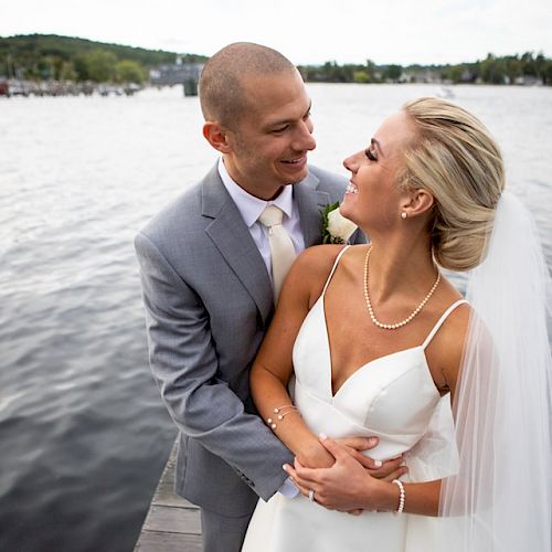 A couple in wedding attire stands on a dock by a lake, looking happily at each other, with greenery in the background.