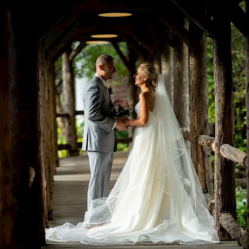 A couple is standing on a wooden bridge, dressed in wedding attire. They are holding hands and looking at each other lovingly, surrounded by nature.