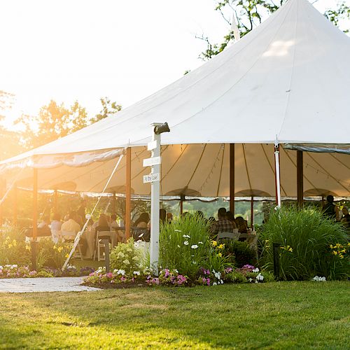 A large white tent set up outdoors, surrounded by lush greenery and flowers, with people seated inside, likely for an event.