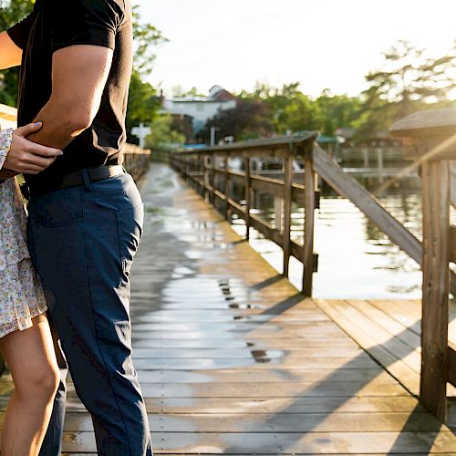 A couple stands on a wooden dock by the water, embracing each other, with greenery and houses in the background during a sunny day.