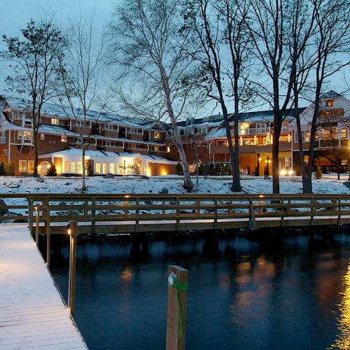 A lit pier leads to a large building by the water, with snow-covered ground and trees, during twilight.