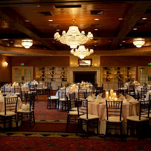 The image depicts an elegant banquet hall with round tables, white tablecloths, dark chairs, and candles on the tables, illuminated by chandeliers.