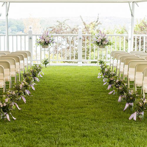 An outdoor wedding setup with rows of white chairs lined with floral decorations on a green lawn, under a white canopy, facing an altar area.