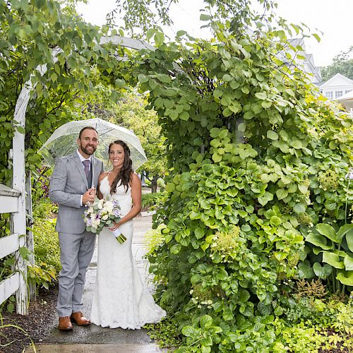 A couple dressed in wedding attire stands under a leafy archway, surrounded by lush greenery and white structures in the background.