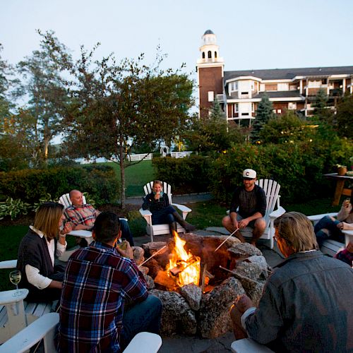 A group of people are sitting around a fire pit in an outdoor area, with a large building in the background.