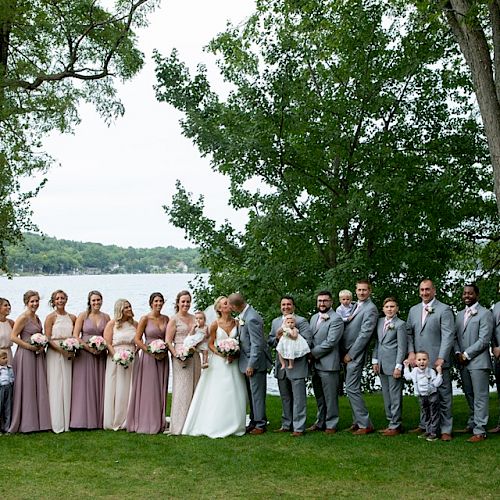 A wedding party stands outdoors in front of a lake and trees. The group includes bridesmaids in lavender dresses and groomsmen in gray suits.