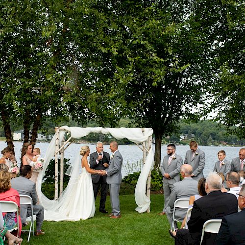 A wedding ceremony is taking place outdoors by a lake with the bride and groom under an arch, surrounded by the bridal party and guests.