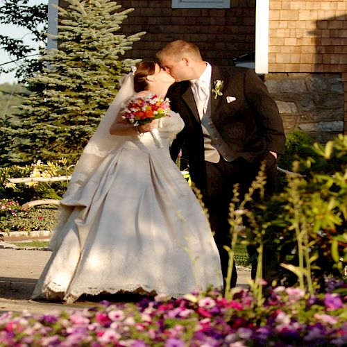 A bride and groom share a kiss in a garden setting, surrounded by lush greenery and vibrant flowers near a wooden building.