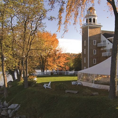 The image shows a scenic riverside view with trees, a large white tent, and a brick building with a clock tower in the background, on a sunny day.