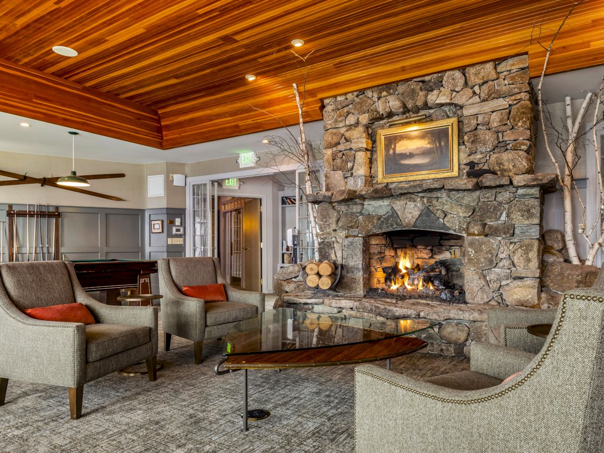 A cozy living room with a stone fireplace, modern furniture, wooden ceiling, and artwork above the mantel. The setting is warm and inviting.