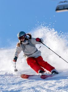 A person is skiing down a snowy slope, wearing a helmet and goggles. A ski lift is visible in the background against a clear blue sky.