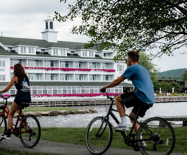 Two people are riding bicycles along a pathway near a large white building with pink flowers on the balcony, next to a body of water.