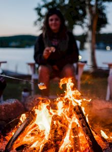 Three people sit around a campfire roasting marshmallows in front of a lake at dusk, enjoying a relaxing evening outdoors.