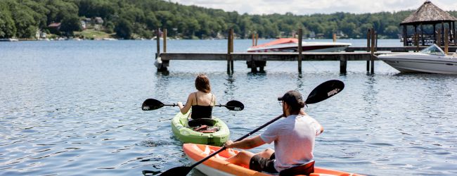 Two people in kayaks paddle on a lake, heading towards a dock with moored boats, surrounded by a wooded shoreline and light clouds overhead.