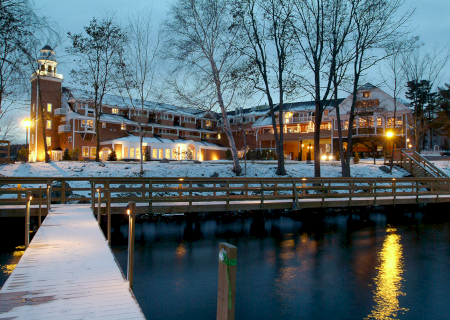 A waterfront building with a lighthouse, surrounded by trees and a wooden dock, under evening light with snow on the ground.