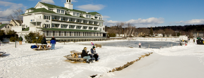 A snowy landscape with people sitting on benches and a large building in the background near a body of water, under a blue sky.