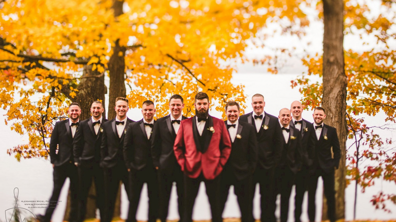 A group of men in formal attire stands in front of autumn trees, with one man in a red jacket at the center, smiling for a photo.