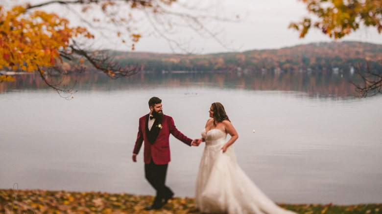 A couple dressed in wedding attire walk hand in hand near a serene lake with autumn foliage surrounding them, creating a romantic atmosphere.