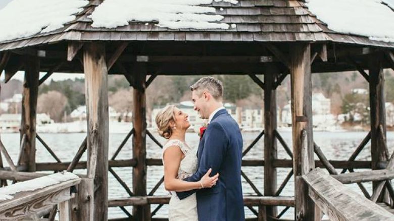 A couple stands on a snowy wooden pier under a gazebo, embracing each other in a serene winter setting.