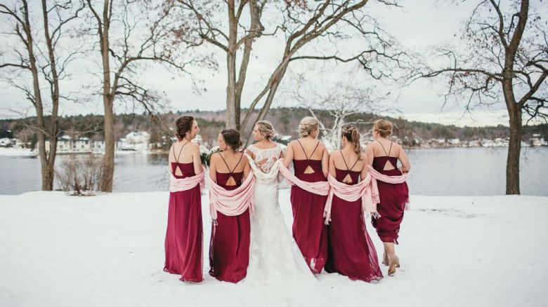 A bride and bridesmaids in burgundy dresses stand together on snowy ground by a lake, with bare trees in the background, posing for a photo.