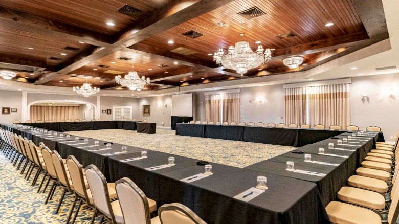 A large conference room with U-shaped seating, tables covered in black cloth, notepads and pens arranged, chandeliers, and a carpeted floor.