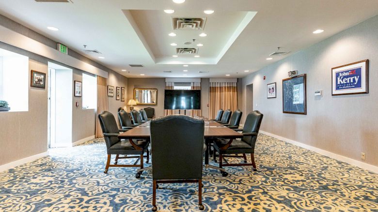 A conference room with a long table, black chairs, framed posters on the wall, ornate blue carpet, and ceiling lights.