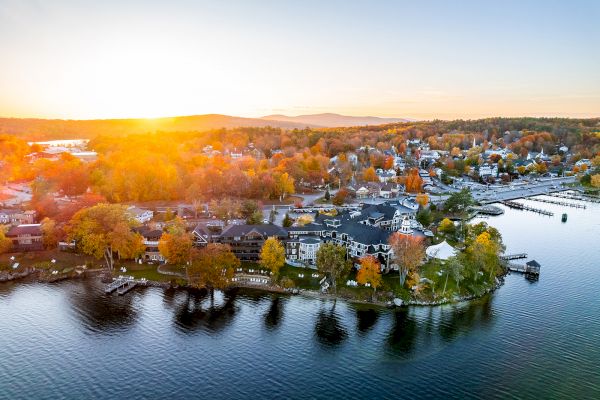 An aerial view of a lakeside town during sunset, showcasing autumn-colored trees, houses, and calm water reflecting the sky.