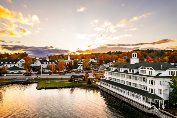 A lakeside town at sunset, featuring a large white building by the shore, colorful autumn foliage, quaint houses, and a scenic waterfront.