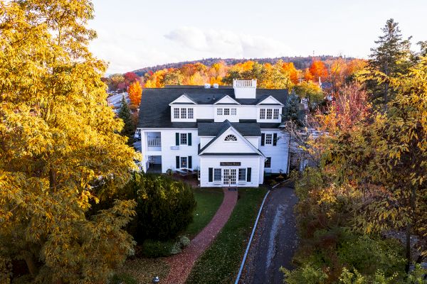 A large, white house surrounded by vibrant autumn foliage with a curved driveway and distant hills in the background.