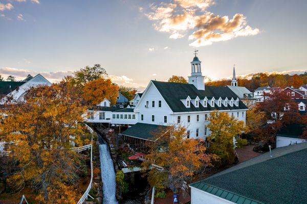 The image shows a picturesque village with white buildings, a church with a steeple, and colorful autumn trees under a partly cloudy sky.