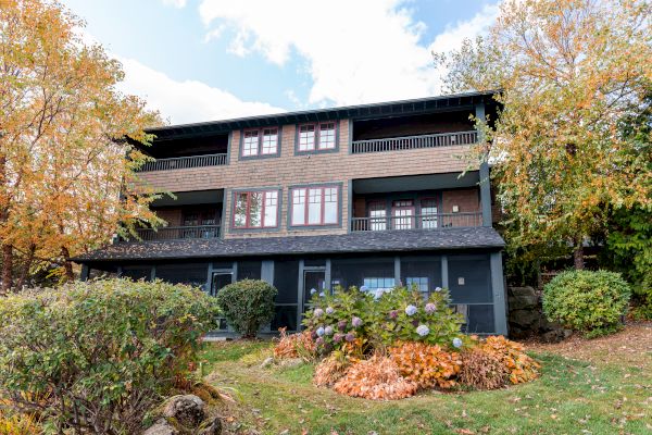 A multi-story house is surrounded by autumn foliage, with colorful plants and a well-maintained yard in the foreground.