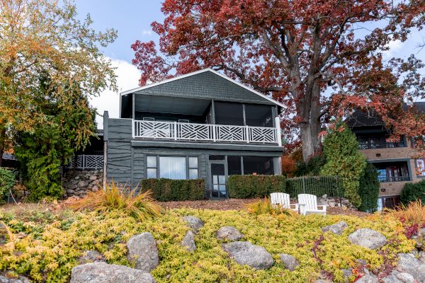 This image shows a two-story house with a porch, surrounded by autumn trees, greenery, rocks, and two white chairs in the garden.