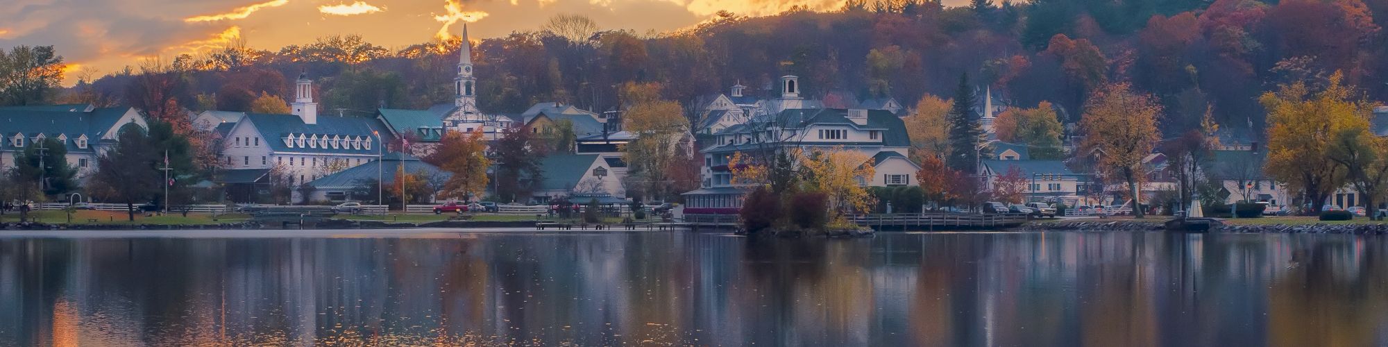 A serene lakeside town at sunset, featuring charming buildings, a church steeple, and colorful autumn trees, reflected in calm water, under a dramatic sky.