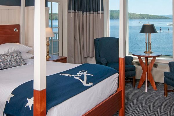 A cozy bedroom with a nautical theme, featuring a four-poster bed, armchairs, and a view of a large body of water through the windows.