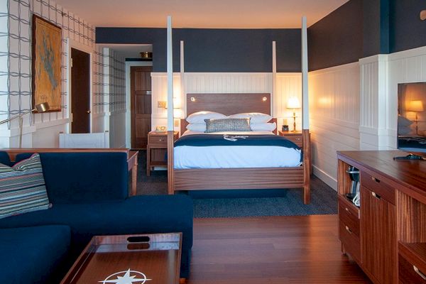 The image shows a cozy hotel room with a four-poster bed, a navy-blue couch, a wooden TV stand, and nautical-themed decor.