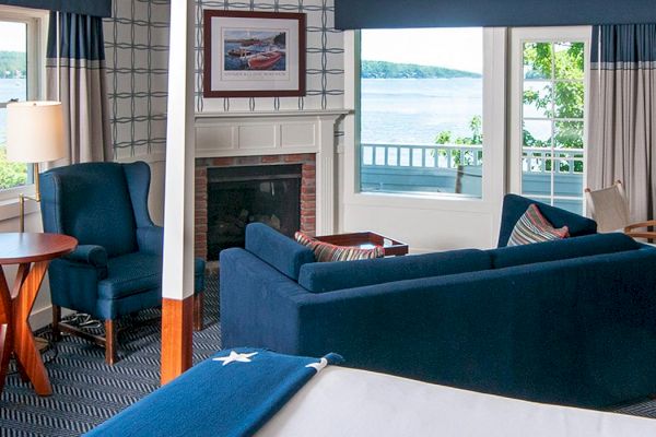 A cozy room with blue furniture, fireplace, and large windows offering a scenic lake view. There's a desk, table, and a bed with a blanket.