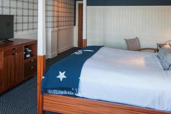 The image shows a neatly arranged hotel room with a large bed, wooden furniture, a TV, and a nightstand with a lamp against a patterned wall.