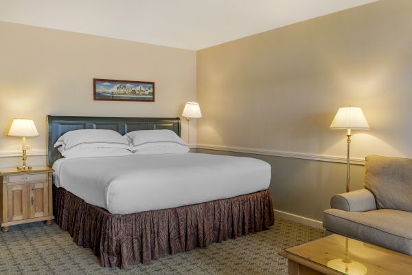 The image shows a cozy hotel room with a neatly made bed, two bedside tables with lamps, a sofa, and a glass-topped coffee table.