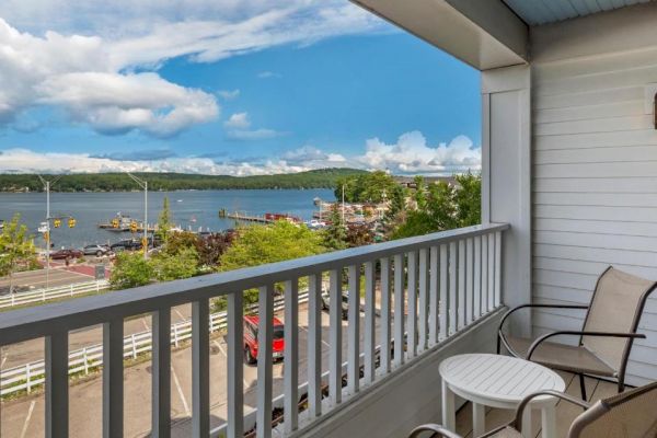 The image shows a balcony with two chairs and a small table, overlooking a scenic view of a lake, green hills, and a few buildings under a partly cloudy sky.