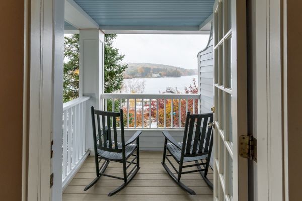 An open door frames a cozy porch with two black rocking chairs facing a scenic view of a lake and autumn trees at a distance.