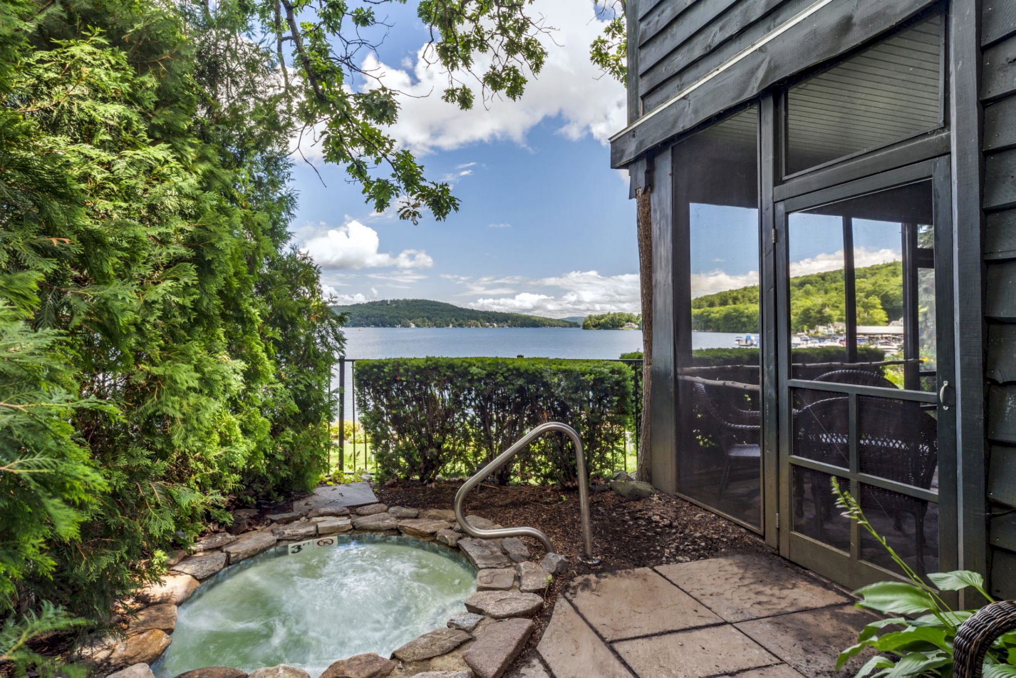 A small outdoor hot tub is surrounded by greenery and stone, with a screened porch and a lake view in the background under a partly cloudy sky.