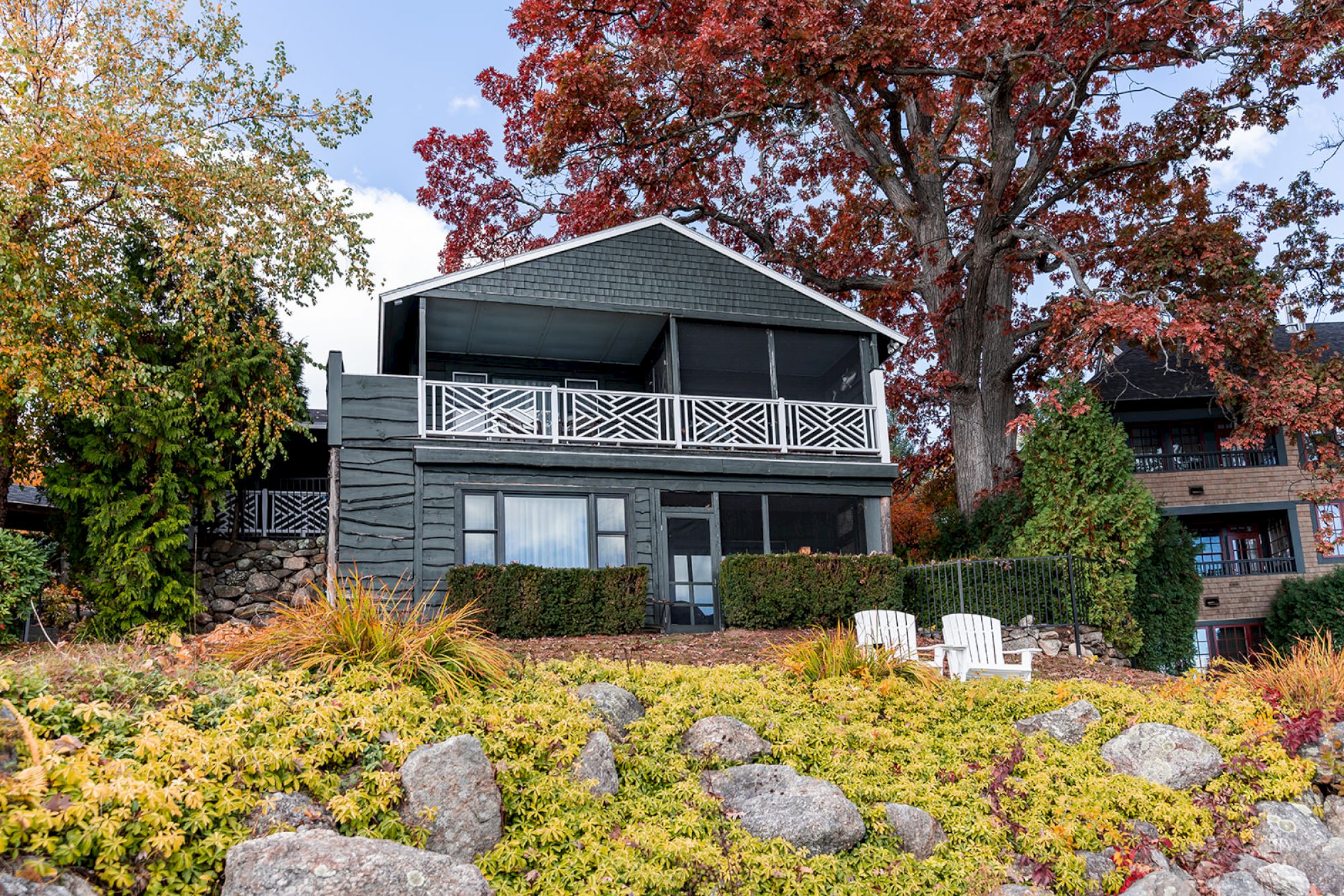 A two-story house features a large upper balcony, surrounded by autumn trees and garden landscaping with rocks and two white chairs.