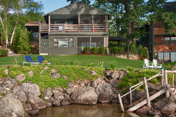 A lakeside house with a large porch, lawn with blue lounge chairs, white chairs, rocks, and a wooden dock beside the clear water's edge.